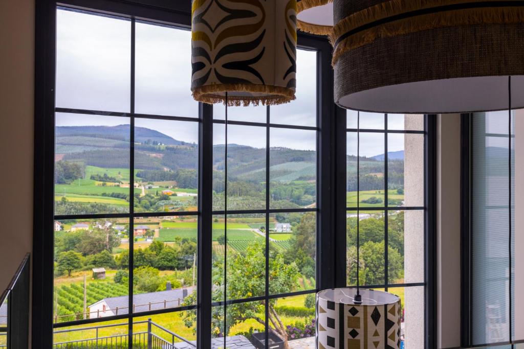 A large window with black frames reveals a scenic view of green fields, scattered houses, and rolling hills—just the kind of landscape you’d expect from boutique hotels in Asturias. Decorative hanging lights are visible inside the room.