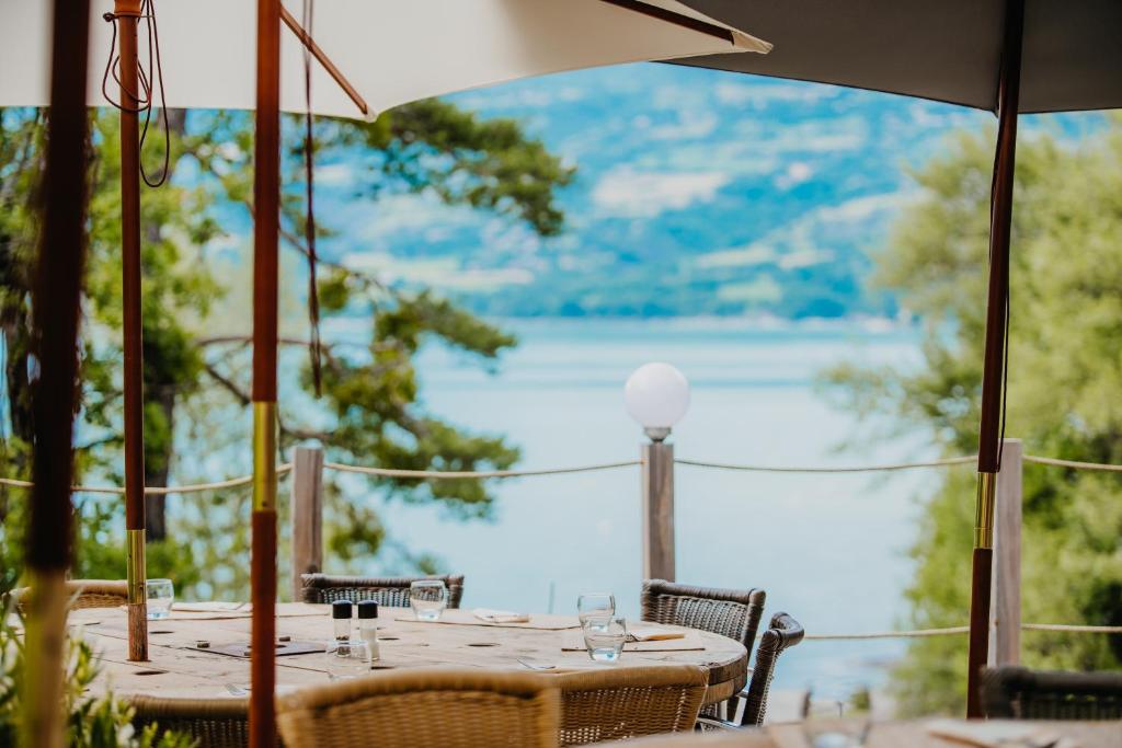11 Unique Places to Stay in Provence 21 Outdoor restaurant seating with round wooden tables and wicker chairs under large umbrellas, overlooking a serene lake surrounded by trees and hills—one of the unique places to stay in Provence. Tables are elegantly set with glasses and cutlery.