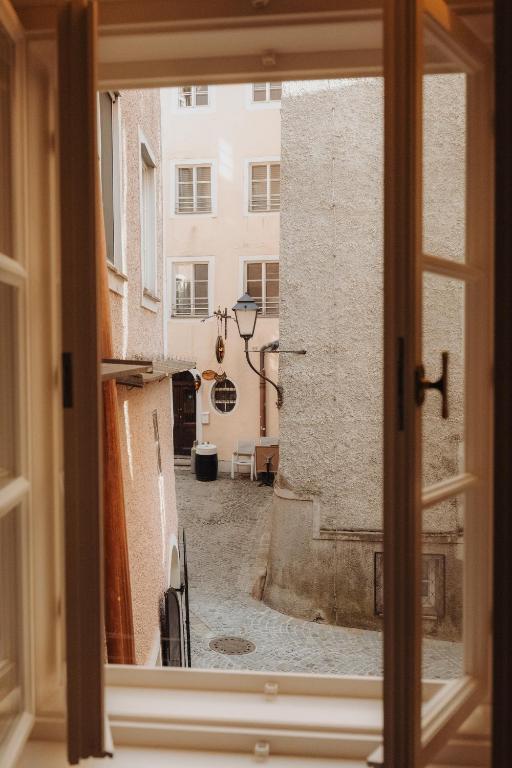 Unique Boutique Hotels in Salzburg 20 Open window view of a narrow cobblestone alley bordered by light-colored buildings, with a streetlamp, round mirror, and a few outdoor chairs—capturing the charm often found near boutique hotels in Salzburg’s quiet, sunlit courtyards.