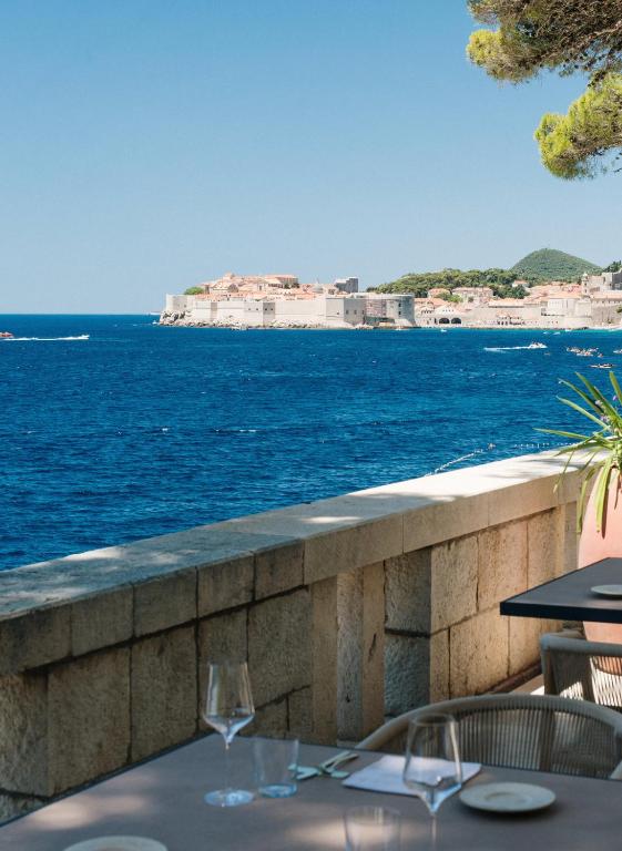 Most Unique Boutique Hotels in Dubrovnik 4 A seaside restaurant terrace overlooks the deep blue water, with stone walls in the foreground and a distant historic town known for its boutique hotels in Dubrovnik, nestled among green hills under a clear blue sky.