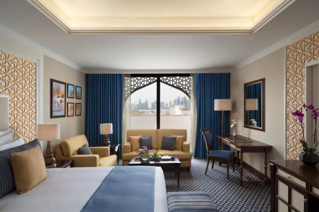 Where to Stay in Doha, Qatar: Transit, City & Beach Hotels 40 Elegant hotel room with a large bed, yellow sofa, armchairs, desk, and lamps—perfect for those seeking where to stay in Doha. Blue curtains frame city views, while patterned walls and framed artwork create a modern, luxurious atmosphere.
