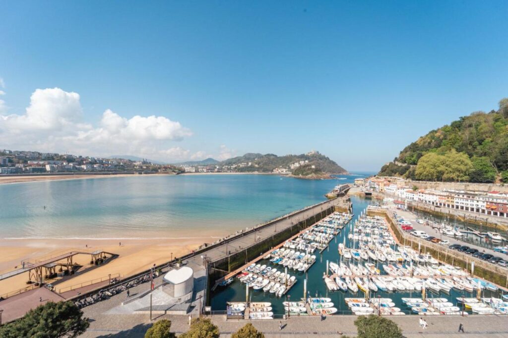 Best Boutique Hotels in San Sebastian 38 A coastal city with boutique hotels in San Sebastian, a marina full of boats, a sandy beach, turquoise water, and green hills under a blue sky. Elegant buildings line the shore while hills rise in the background.