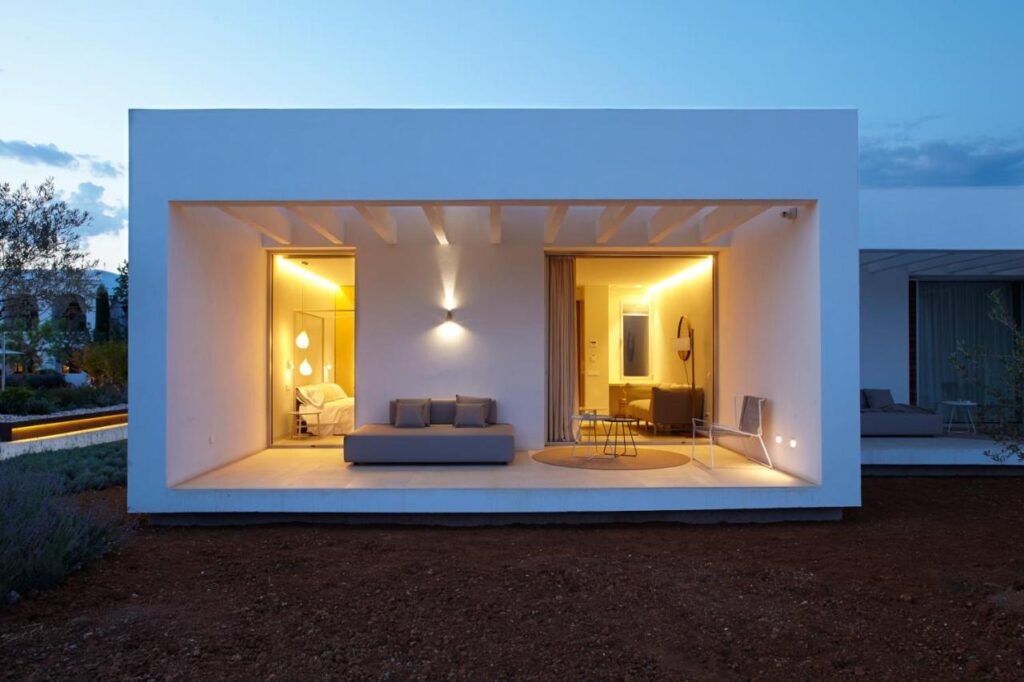 10 AUTHENTIC Finca Hotels in Ibiza 36 Modern white house with large open patio, warm yellow lighting inside, visible living area with sofa, and a bedroom to the left. The outdoor area has bare soil and greenery, reminiscent of finca hotels in Ibiza under an evening sky.