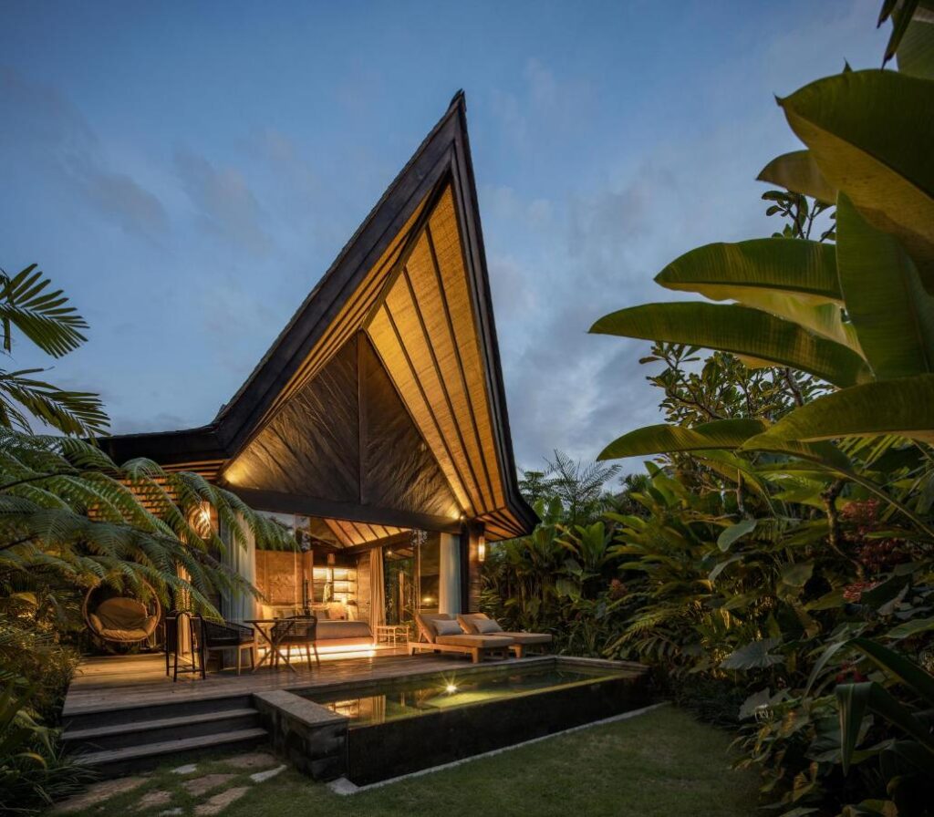 14 UNIQUE Boutique Hotels in Canggu, Bali 8 A modern tropical villa with a steep, triangular wooden roof sits among lush greenery at dusk, resembling the inviting charm of boutique hotels in Canggu, featuring open living, warm lighting, outdoor seating, and a small rectangular pool in front.
