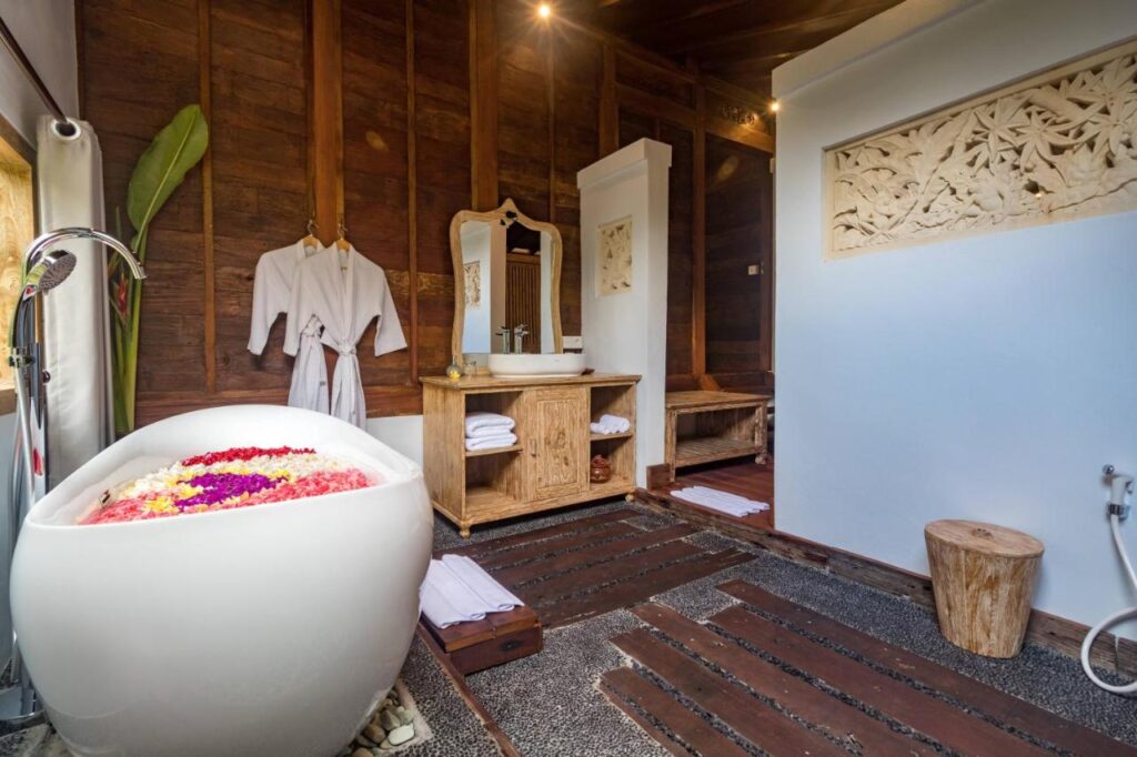 13 Most UNIQUE Places to Stay in Ubud 25 A luxurious bathroom with wooden walls, a white freestanding bathtub filled with flower petals, two white robes hanging by a mirror above a wooden vanity, and a carved wall decoration—perfect for those seeking unique places to stay in Ubud.