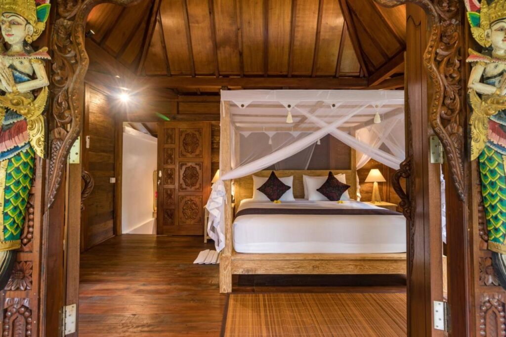 13 Most UNIQUE Places to Stay in Ubud 23 A cozy bedroom with carved wooden decor, a large canopy bed with white netting, colorful pillows, warm lighting, and intricate statues framing the entrance—one of the unique places to stay in Ubud with rustic, tropical, and elegant design.