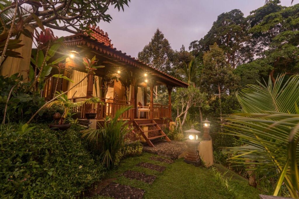 13 Most UNIQUE Places to Stay in Ubud 22 A cozy wooden cottage with a tiled roof is lit by warm lights, surrounded by lush greenery and tropical plants—a perfect example of unique places to stay in Ubud, with a stone path leading to the front steps at dusk.