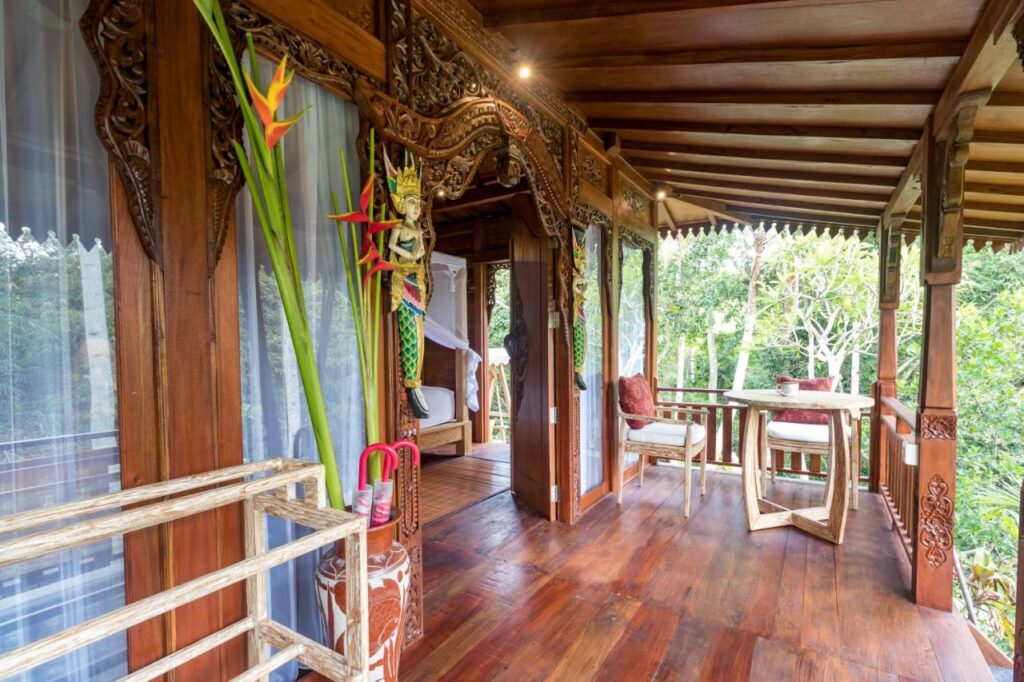 13 Most UNIQUE Places to Stay in Ubud 24 A wooden balcony with ornate carvings, large windows with curtains, tropical plants, and a small table with chairs overlooks lush green trees in a serene outdoor setting—one of the unique places to stay in Ubud.