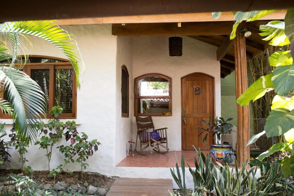 Unique Hotels in Costa Rica: Top Stays 6 A cozy house entrance with a wooden door, arched windows, potted plants, and a wooden rocking chair on a small tiled porch, surrounded by lush green foliage—reminiscent of the charm found in unique hotels in Costa Rica.