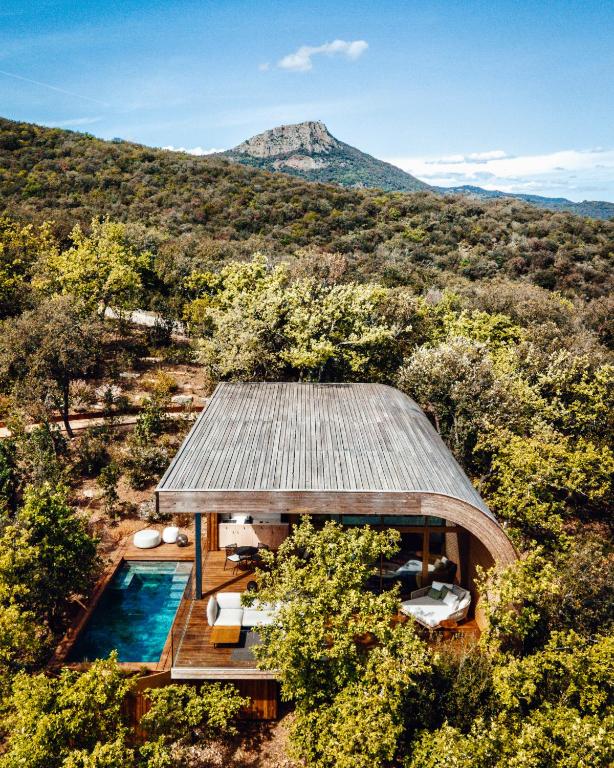 11 Unique Places to Stay in Provence 25 A modern wooden villa with a curved roof sits among dense green trees, featuring an outdoor pool and lounge area. One of the unique places to stay in Provence, it boasts mountain views under a bright blue sky with a few clouds.