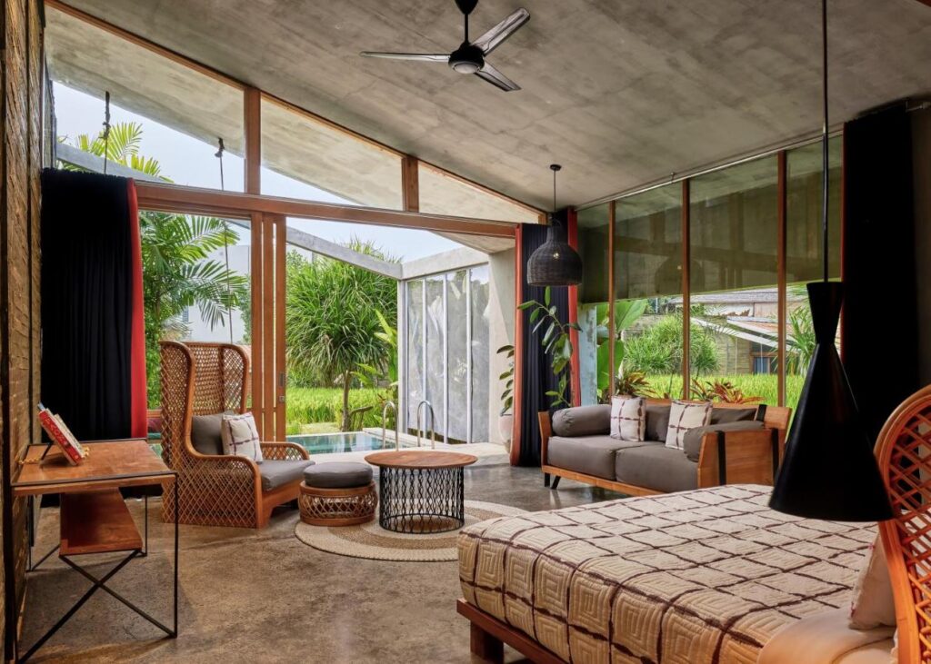 14 UNIQUE Boutique Hotels in Canggu, Bali 21 A modern tropical living space inspired by boutique hotels in Canggu, with wooden furniture, large glass windows, a ceiling fan, woven chairs, and views of a private pool and lush greenery that create an open, airy atmosphere.