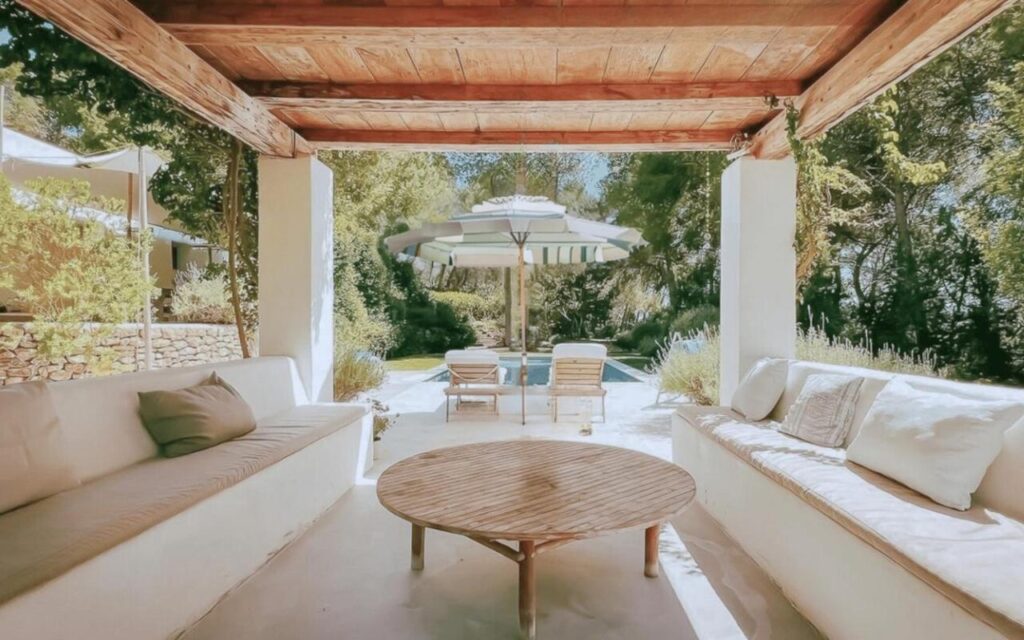 10 AUTHENTIC Finca Hotels in Ibiza 38 A covered patio with cushioned benches and a round wooden table overlooks a sunlit pool area with lounge chairs and a striped umbrella, surrounded by greenery—evoking the relaxed charm of finca hotels in Ibiza.