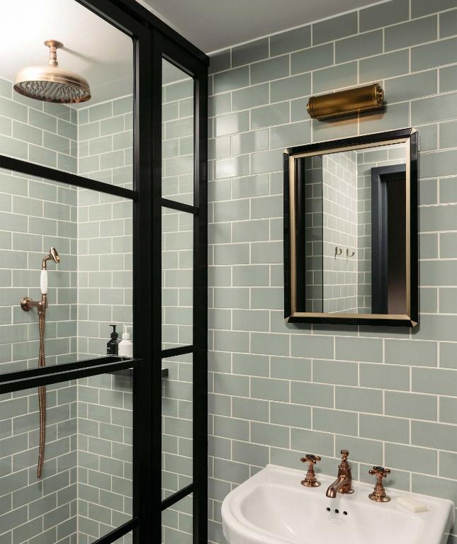 15 Best Boutique Hotels in London, UK 59 Modern bathroom with light green subway tiles, a glass shower enclosure with black frames—reminiscent of boutique hotels in London—features a rainfall showerhead, white sink with bronze fixtures, rectangular mirror, and brass wall light above.