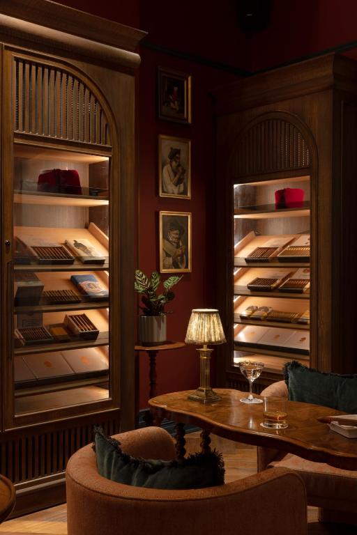 15 Best Boutique Hotels in London, UK 32 A warmly lit lounge in the style of boutique hotels in London, with wooden cigar cabinets, velvet chairs, a small table with drinks, a vintage lamp, and framed art on deep red walls, creating an elegant and cozy atmosphere.