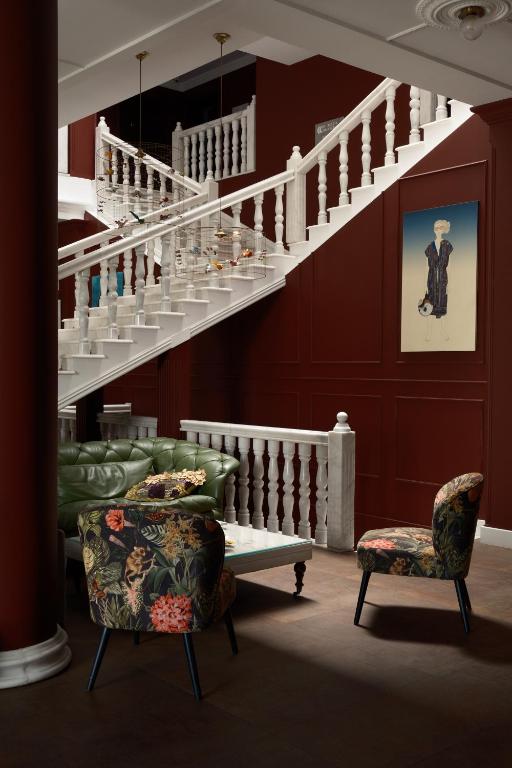 11 Unique Hotels in Georgia You Can't Miss! 25 A stylish interior at one of the unique hotels in Georgia features a green tufted sofa, floral-patterned chairs, and a white coffee table beneath a grand staircase set against dark red walls adorned with modern art.