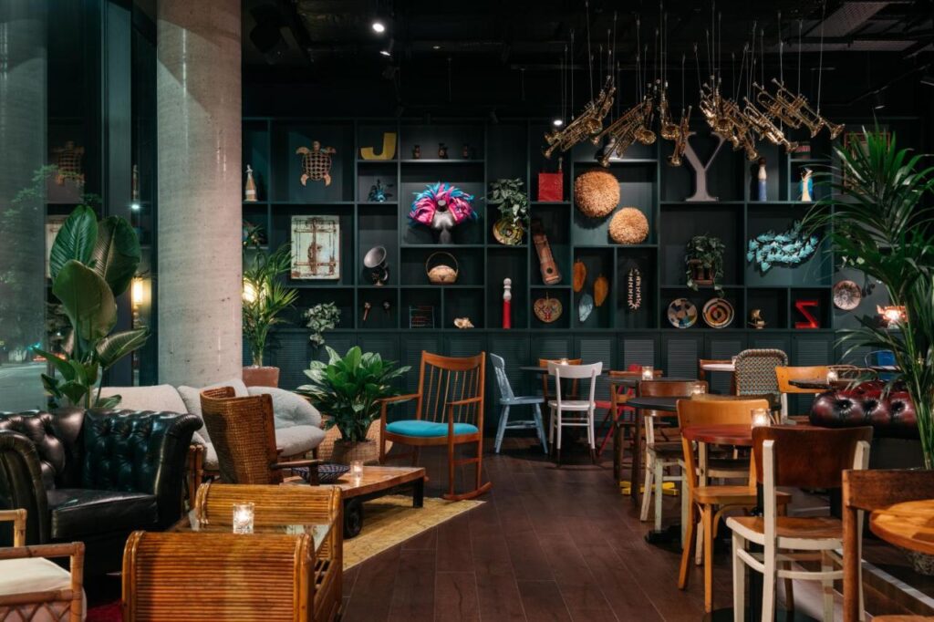 15 Best Boutique Hotels in London, UK 18 A stylish, eclectic café reminiscent of the best boutique hotels London offers, featuring various chairs, wooden tables, lush plants, and a dark shelving unit displaying art and books in a cozy, warmly lit atmosphere.