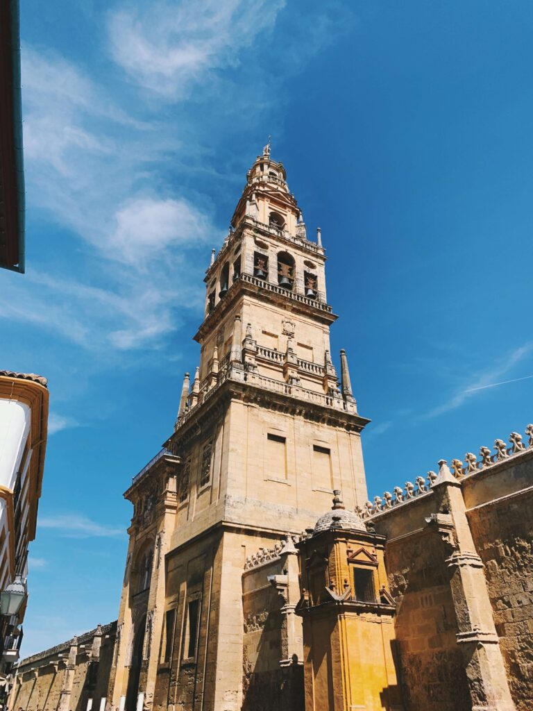 Best of Cordoba One Day Itinerary 11 A tall, ornate stone bell tower rises against a bright blue sky in Cordoba, attached to a historic building with detailed architectural features and arched windows—perfect for exploring on a Cordoba one day visit.
