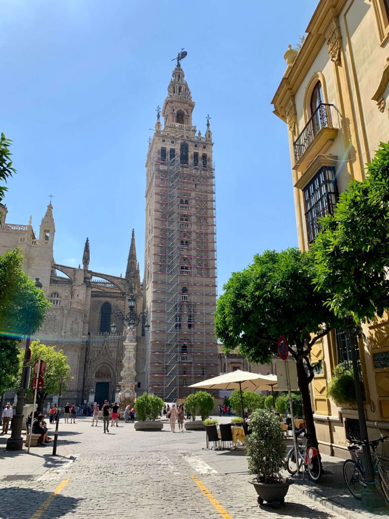 1 day in Seville Spain: My Favorite City 18 The Giralda tower in Seville, Spain, rises above a sunny plaza with scaffolding along its facade—an iconic sight for anyone spending 1 day in Seville, Spain—surrounded by historic buildings, trees, people, and outdoor seating.