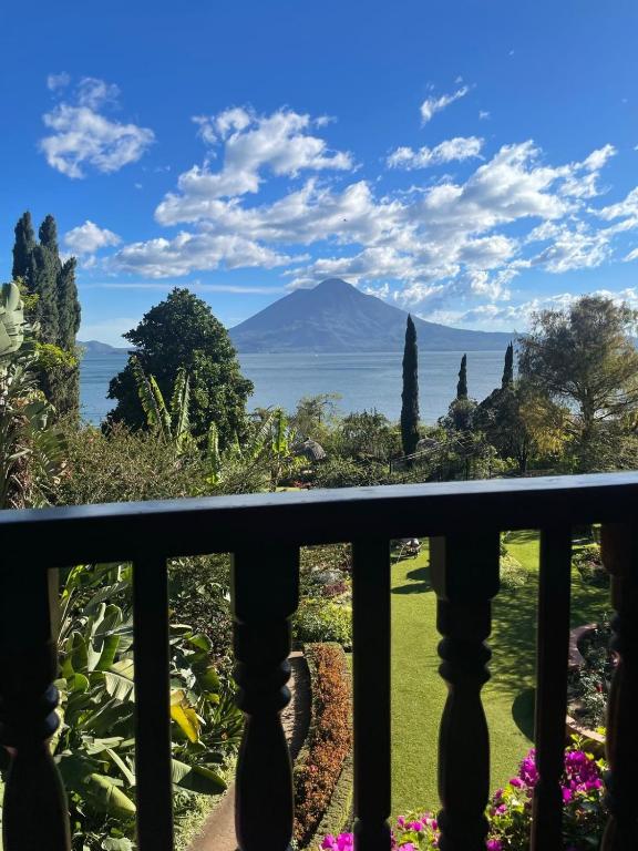 8 UNIQUE Places Where to Stay in Lake Atitlán 30 A scenic view of a lush garden with colorful flowers, tall trees, and a lake, framed by a wooden balcony railing—perfect inspiration for those seeking where to stay in Lake Atitlan, with mountains and a partly cloudy blue sky in the background.