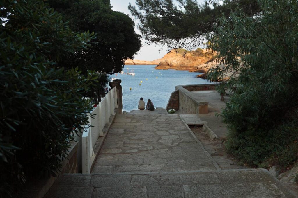 A stone pathway leads down to a calm seaside near boutique hotels in Costa Brava, with two people sitting by the water’s edge. Trees and bushes frame the path, and yellow buoys float in the blue water under a clear sky.
