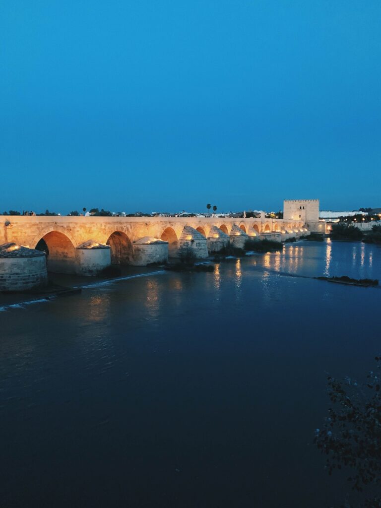 Best of Cordoba One Day Itinerary 8 A historic stone bridge with arches is illuminated by golden lights at dusk, reflecting on a calm river, with a large square tower at one end—an unforgettable Cordoba one day scene beneath the deep blue sky.