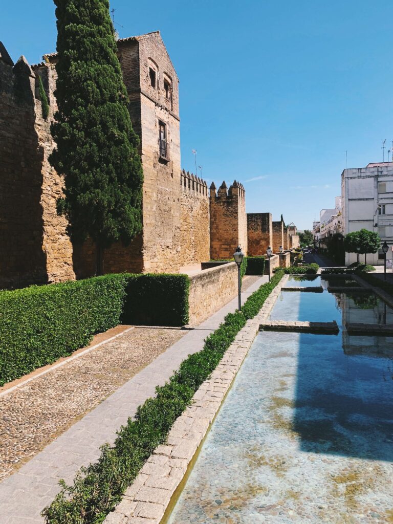 Best of Cordoba One Day Itinerary 6 A stone walkway with hedges runs beside a clear reflecting pool and an old fortress wall—a picturesque scene perfect for exploring Cordoba one day, with tall cypress trees and modern white buildings under a blue sky.