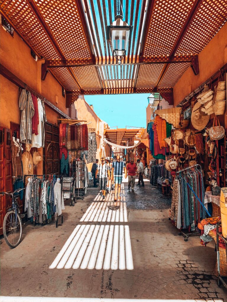 10 Unique Riads in Marrakech with Pool 43 A lively outdoor market with orange walls and a wooden slatted roof, near budget riads in Marrakech. Colorful clothes, hats, and baskets hang on both sides as people stroll the sunlit path; a bicycle rests against the left wall.