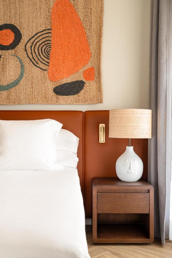 10 UNIQUE Hotels with Pool in Seville 29 A neatly made bed with white linens sits next to a wooden nightstand holding a white lamp with a woven shade, reminiscent of the cozy style found in hotels with pool in Seville. Above the bed hangs abstract textile art in warm tones; gray curtains frame the scene.