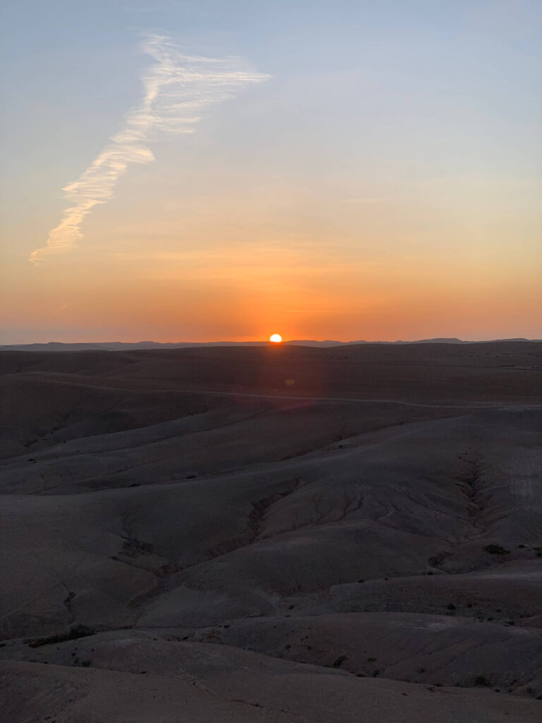 8 SPECTACULAR Agafay Desert Camps 7 Sunset over a vast, barren desert landscape with rolling hills in the foreground and a streak of cloud in the sky, casting a warm orange and yellow glow near Agafay desert camps on the horizon.
