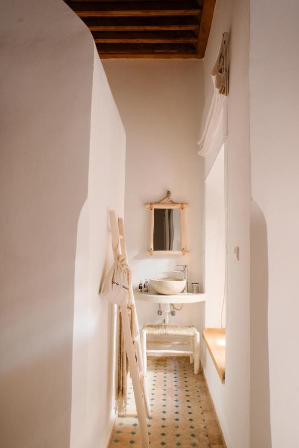 12 Charming Budget Riads in Marrakech 53 A narrow bathroom in the style of budget riads in Marrakech, featuring white walls, a small sink with a mirror above, wooden ceiling beams, patterned floor tiles, and a tote bag by the entrance. Natural light streams through a window above a bench.
