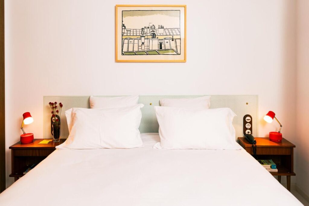 Where to Stay in Madrid: 14 Best Hotels 41 A neatly made bed with white pillows and sheets sits between two wooden nightstands, each with a red lamp—a cozy spot for those wondering where to stay in Madrid first time. A framed drawing of rooftops hangs on the white wall above the bed.
