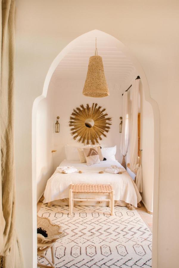 12 Charming Budget Riads in Marrakech 50 A cozy bedroom inspired by budget riads in Marrakech, featuring a white arched doorway, sunburst mirror above the bed, woven pendant light, and natural wood accents in a warm, neutral color palette.