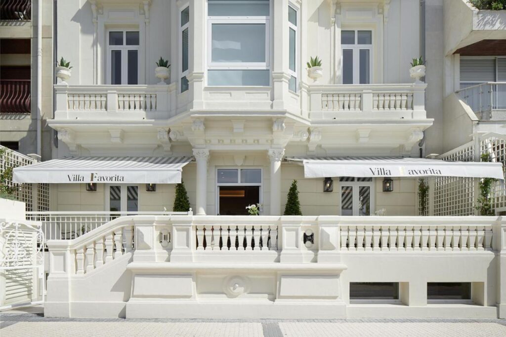 Best Boutique Hotels in San Sebastian 19 Elegant white building with decorative columns, balconies, and striped awnings that read Villa Favorita—one of the top boutique hotels in San Sebastian. Potted plants and a balustrade line the front, with steps leading to the main door.