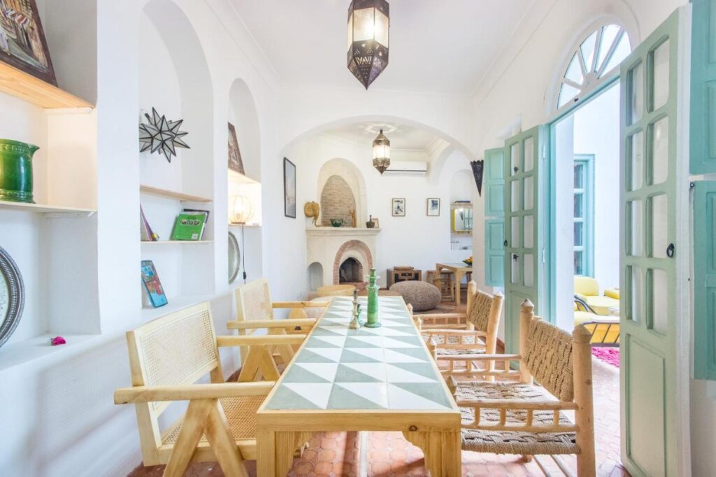 12 Charming Budget Riads in Marrakech 19 Bright, airy room with a long table and wooden chairs, geometric patterned tabletop, arched doorways, and open double doors—reminiscent of the inviting charm found in budget riads in Marrakech—decorated in minimalist Mediterranean style.