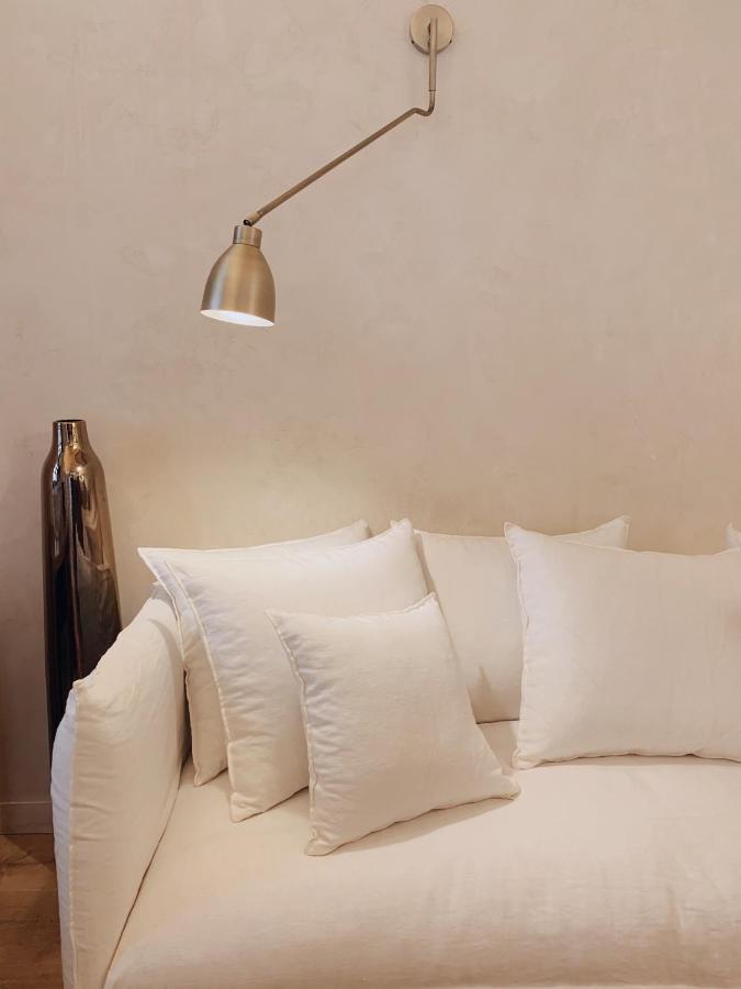 A modern, minimalist living room—reminiscent of boutique hotels in Costa Brava—features a white sofa with matching cushions, a metallic wall-mounted reading lamp above, and a tall, shiny vase set against a light beige wall.