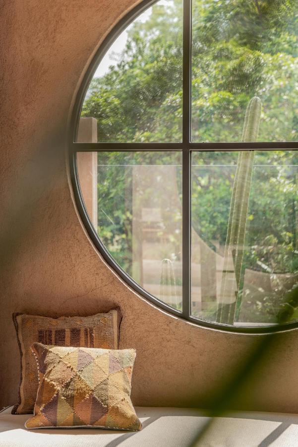 A cozy seating area with two patterned cushions sits by a large circular window, revealing green plants and trees outside on a sunny day—an inviting escape found in the best family villas in Bali. Soft natural light fills the earthy-toned room.