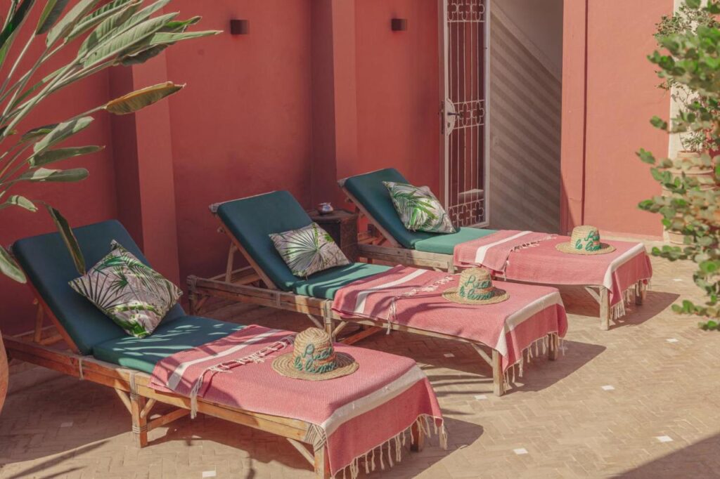 12 Charming Budget Riads in Marrakech 30 Three wooden lounge chairs with green cushions and tropical-patterned pillows sit against a pink wall, evoking the relaxed charm of budget riads in Marrakech. Each chair is topped with a straw hat and pink towel, surrounded by leafy plants.