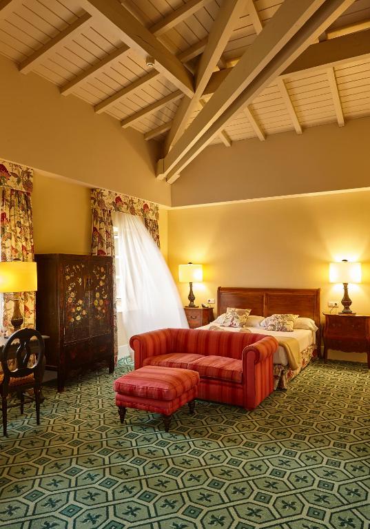 8 Unique Boutique Hotels in Bilbao Spain 20 A cozy bedroom inspired by boutique hotels in Bilbao, featuring a wooden beamed ceiling, patterned carpet, floral curtains, a wooden-framed bed, ornate armoire, bedside tables with lamps, and a red-striped sofa and ottoman at the foot of the bed.