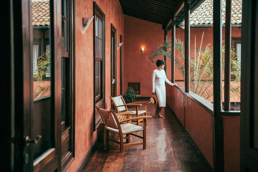 11 UNIQUE Boutique Hotels in Tenerife 35 A person in a white dress stands on a wooden balcony with terracotta walls, holding a hat and gazing at tropical plants—a serene scene often found at boutique hotels in Tenerife. Two wooden chairs and a small table complete the tranquil setting.