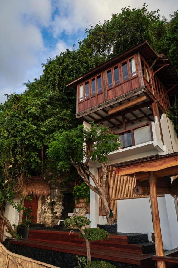 Nestled against a lush, green hillside, this multi-level wooden and white building with large windows and surrounding trees is one of the best family villas in Bali, blending natural materials for a serene, tropical atmosphere.