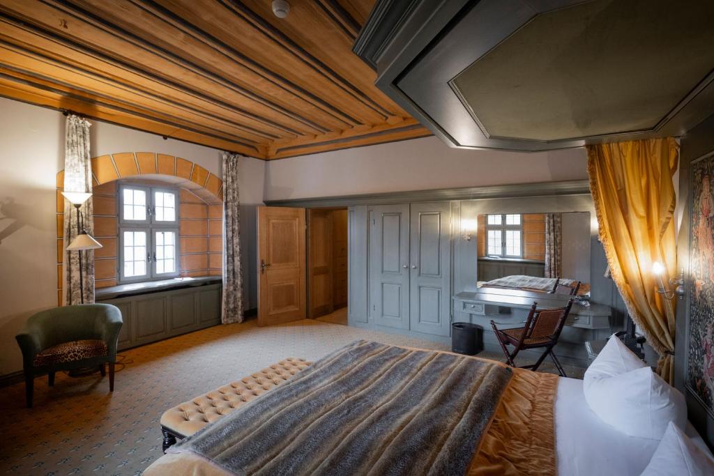 FAIRYTALE Castle Hotels in Bavaria 14 A spacious, elegantly decorated hotel room reminiscent of castle hotels in Bavaria, featuring a wooden ceiling, large window, double bed with gray and gold bedding, an armchair, built-in wardrobe with mirror and chair, all bathed in warm natural light.