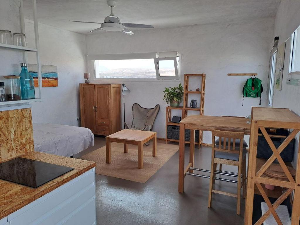 Adults Only Hotels in Fuerteventura 13 A bright, minimalist studio with white walls, wooden furniture, a bed, table, shelves, wardrobe, ceiling fan, and a window offering views of distant hills—ideal for those seeking the tranquility of adults only hotels in Fuerteventura. Plants and simple decor add coziness.