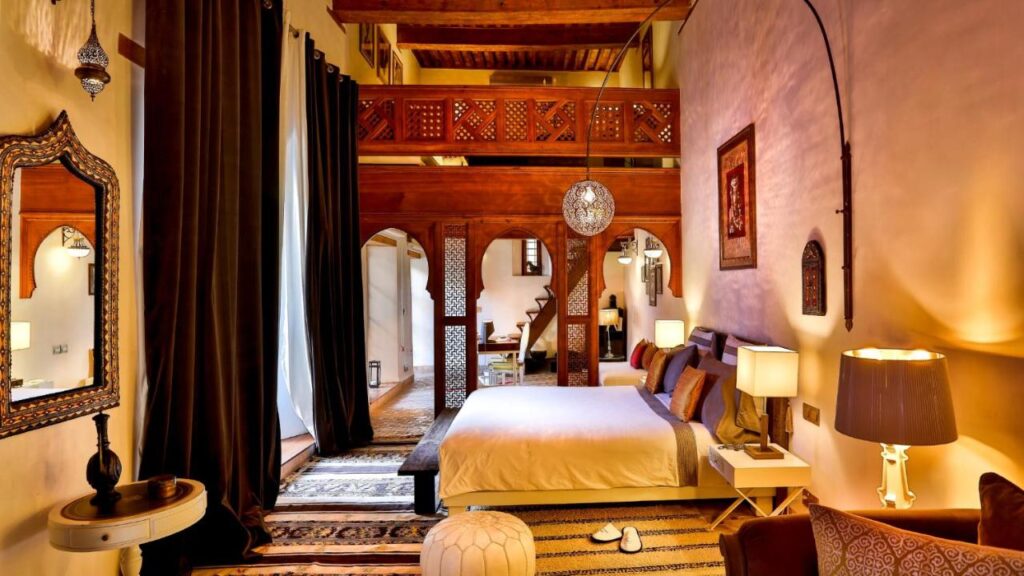 7 UNIQUE Riads in Fez You Won't Forget 8 A cozy bedroom inspired by riads in Fez, featuring Moroccan decor with carved wooden balconies, patterned rugs, a double bed with pillows, hanging lanterns, and warm lighting. Stairs lead to a charming loft area above the bed.