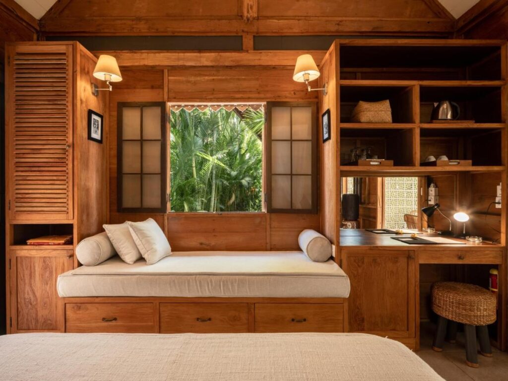 14 UNIQUE Boutique Hotels in Canggu, Bali 44 Cozy wooden room with a built-in daybed below a window, surrounded by open shelves and a desk—reminiscent of boutique hotels in Canggu. Two wall lamps are above the bed, and tropical greenery is visible through the window.