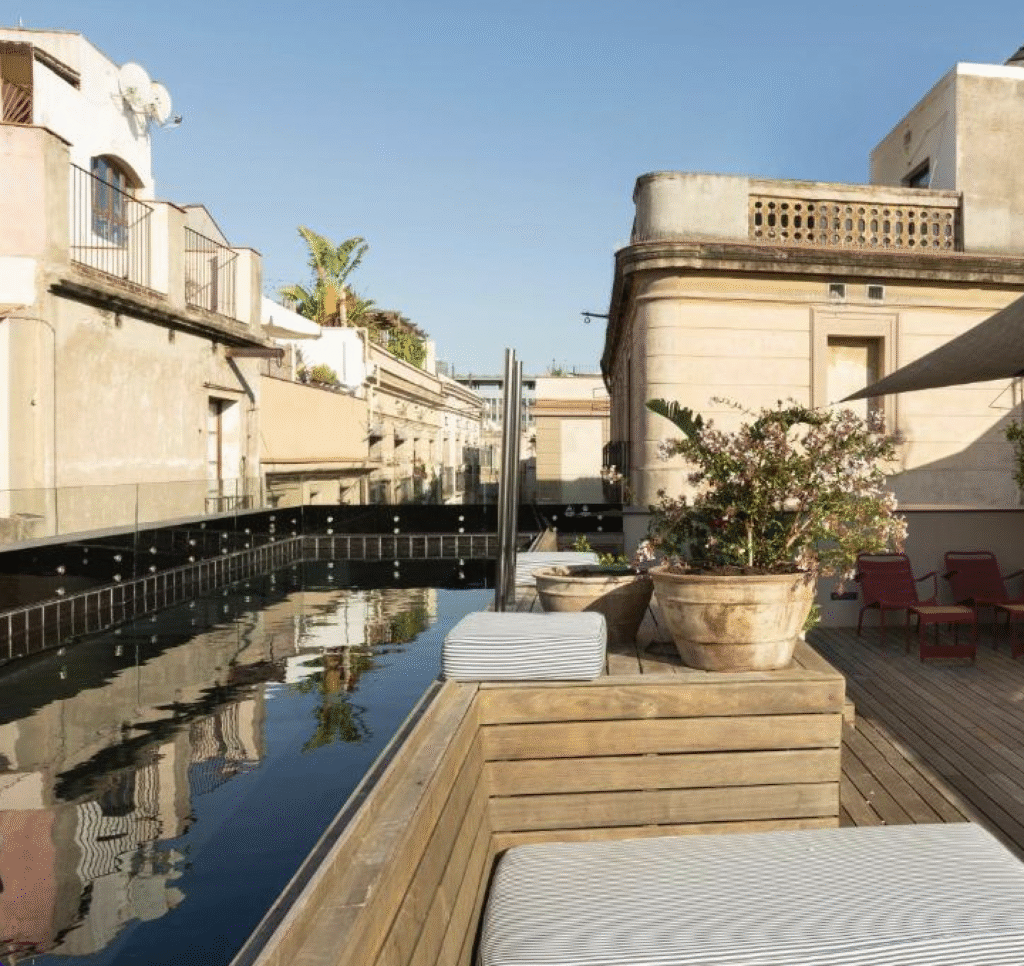 13 Unique Hotels in Barcelona, Spain 42 A rooftop terrace with cushioned wooden seating, potted plants, and a reflective narrow pool evokes the charm of Barcelona hotels with rooftop pool, surrounded by old buildings under a clear blue sky.
