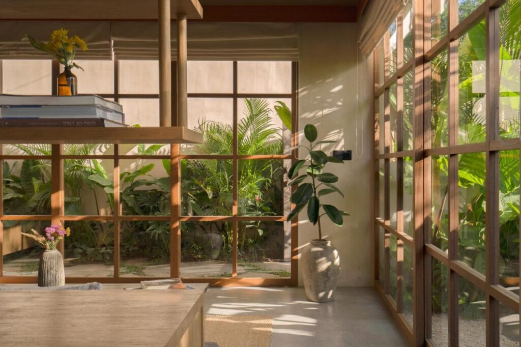 Sunlit room with large glass windows and wooden frames, filled with natural light. Like the best family villas in Bali, lush green plants thrive inside and out. A table holds stacked books and flower vases, creating a cozy, natural atmosphere.