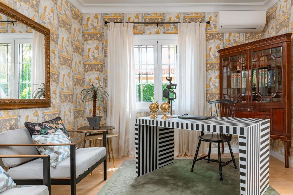 11 UNIQUE Boutique Hotels in Tenerife 44 A stylish home office inspired by boutique hotels in Tenerife, featuring patterned wallpaper, a black-and-white striped desk, a black chair, a glass-front cabinet, a sofa with patterned pillows, a potted plant, and large windows with sheer curtains.