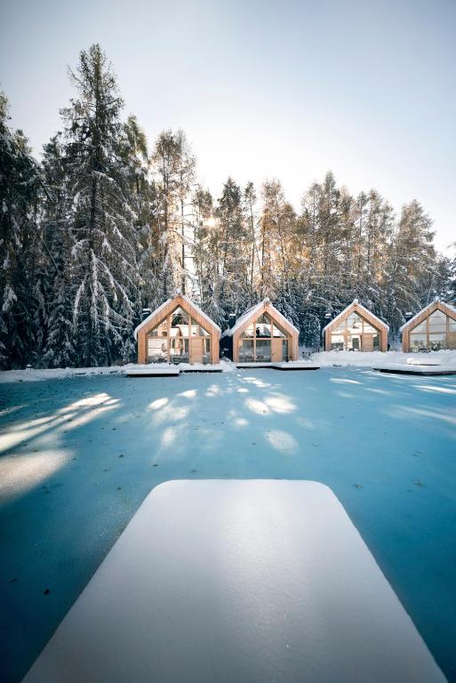 Four small wooden cabins with large windows stand in a snowy forest. In the foreground, a snow-covered surface and a frozen blue lake evoke the serene beauty found at some of the best hotels in the Dolomites, surrounded by tall pine trees.
