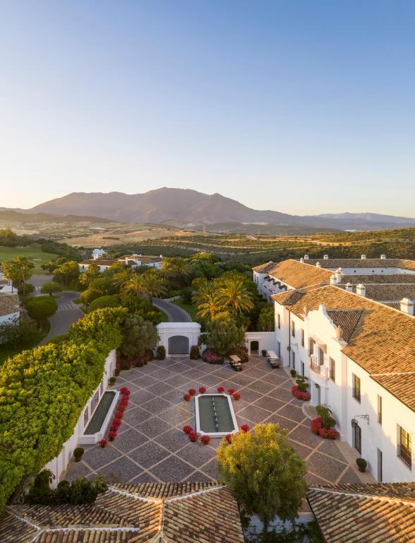 TOP 10 Luxury Hotels in Andalucía 6 Aerial view of a Mediterranean-style courtyard at one of the luxury hotels in Andalucía, featuring a central fountain, white buildings, lush greenery, and mountains in the background under a clear blue sky.