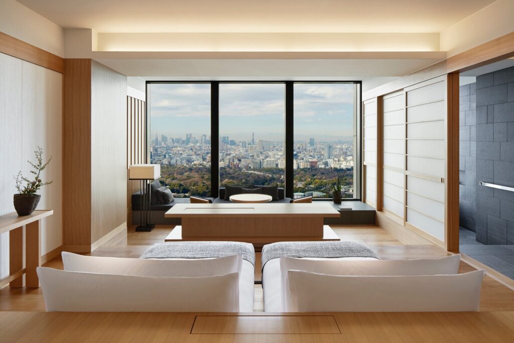 15 Unique Hotels in Tokyo: Coolest hotels 26 A modern, minimalist hotel room with light wood accents, white furnishings, and large windows offering a panoramic city view—an inviting space that stands out among unique hotels in Tokyo. A low table by the window and a sliding door lead to a private bath area.